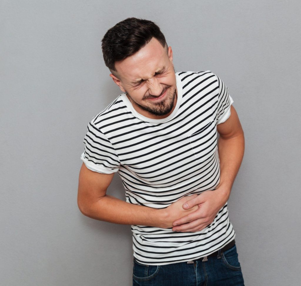 Image of displeased young man with painful feelings holding his belly isolated over grey background. Eyes closed.